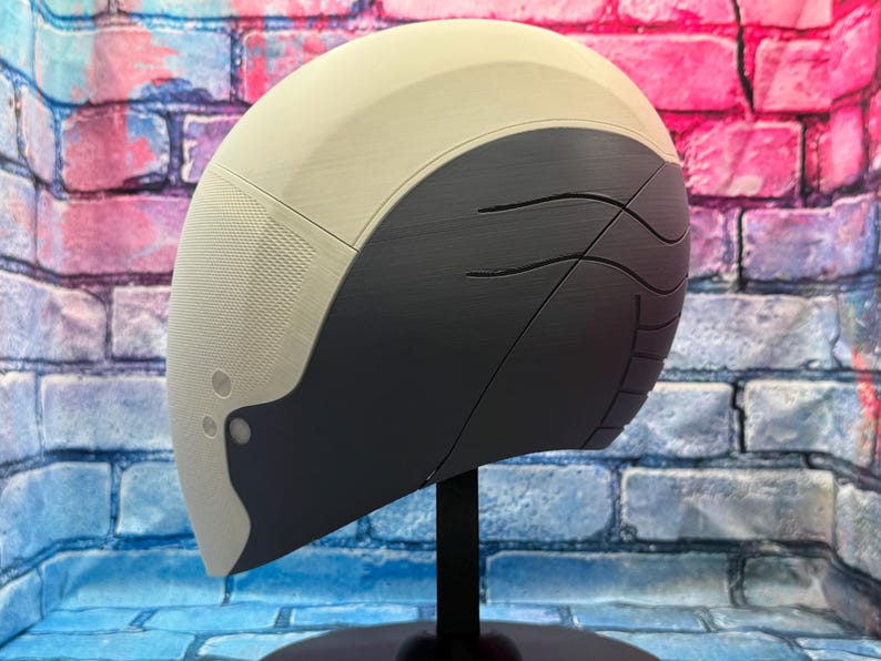 May include: A futuristic-looking helmet with a two-tone design. The top portion is off-white, while the lower section is dark gray with curved etched lines. The helmet is mounted on a black stand, set against a brick wall.