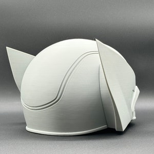 Puede incluir: Un casco blanco impreso en 3D con una parte superior redondeada y dos piezas de oreja puntiagudas. El casco tiene un acabado liso y mate.
