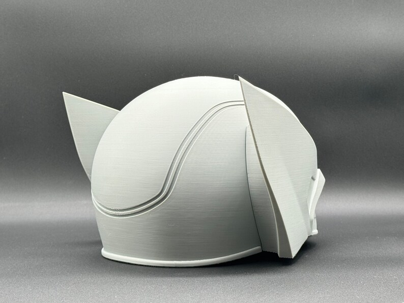 Puede incluir: Un casco blanco impreso en 3D con una parte superior redondeada y dos piezas de oreja puntiagudas. El casco tiene un acabado liso y mate.