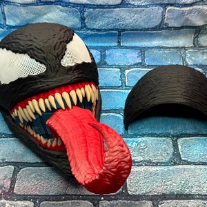 May include: A black Venom mask with white mesh eyes, sharp white teeth, and a long red tongue. The mask has a textured surface and is accompanied by a separate black headpiece. The mask is a cosplay item.