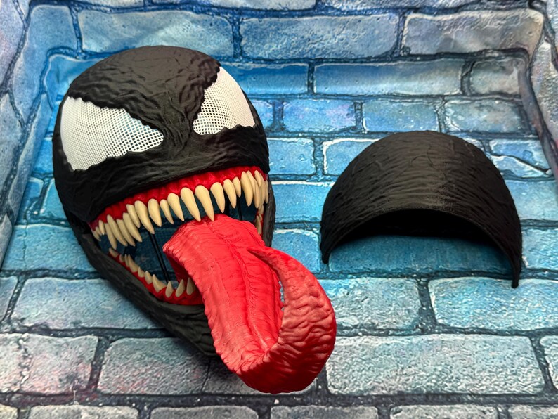 May include: A black Venom mask with white mesh eyes, sharp white teeth, and a long red tongue. The mask has a textured surface and is accompanied by a separate black headpiece. The mask is a cosplay item.