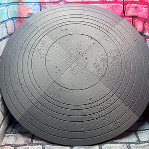 May include: A gray, circular shield with concentric rings and a central cross design. The shield has a textured surface and appears to be made of metal. The background features a brick wall with colorful graffiti.