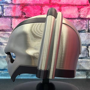 May include: A silver and black helmet with a unique design, featuring a visor and side attachments. The helmet is displayed against a colorful brick wall background.