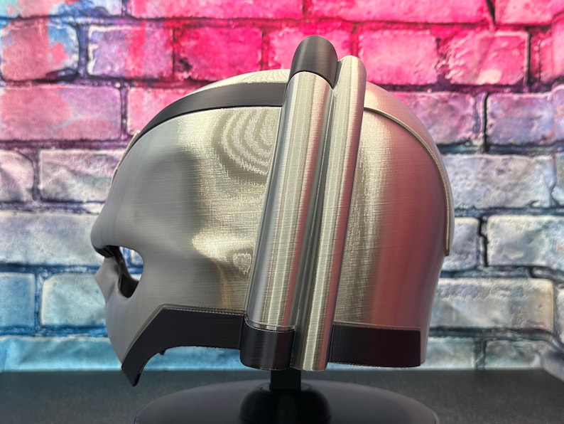 May include: A silver and black helmet with a unique design, featuring a visor and side attachments. The helmet is displayed against a colorful brick wall background.