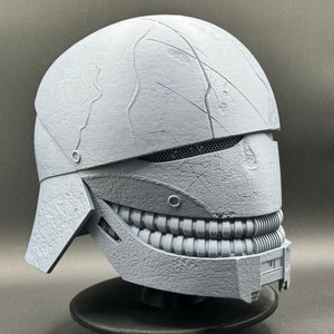 May include: A gray 3D printed helmet with a textured surface and a toothy grin. The helmet has a visor with a mesh design and a small, round detail on the side.