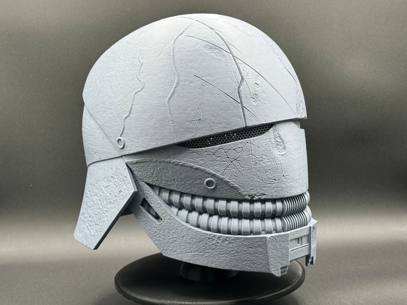 May include: A gray 3D printed helmet with a textured surface and a toothy grin. The helmet has a visor with a mesh design and a small, round detail on the side.