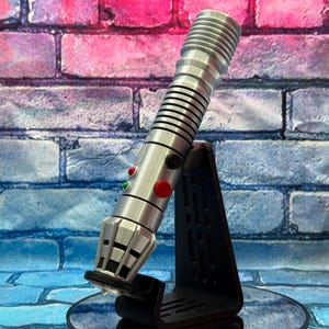 May include: A silver lightsaber with black accents, featuring red and green buttons, is displayed on a black stand. The lightsaber has a ribbed handle and a detailed emitter. The background is a brick wall with pink and blue hues.