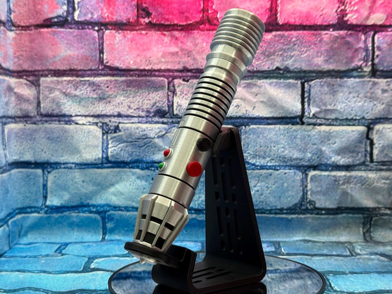 May include: A silver lightsaber with black accents, featuring red and green buttons, is displayed on a black stand. The lightsaber has a ribbed handle and a detailed emitter. The background is a brick wall with pink and blue hues.
