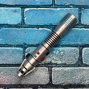 May include: A silver lightsaber prop with black and red buttons, and black stripes. The lightsaber is set against a blue brick wall background. The prop has a detailed design and is likely a collectible item.