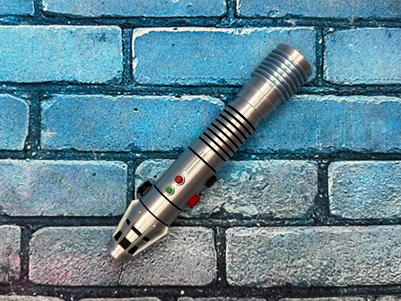 May include: A silver lightsaber prop with black and red buttons, and black stripes. The lightsaber is set against a blue brick wall background. The prop has a detailed design and is likely a collectible item.