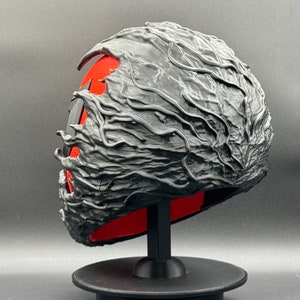 Puede incluir: Una m&aacute;scara negra impresa en 3D con un dise&ntilde;o texturizado y org&aacute;nico. La m&aacute;scara est&aacute; en un soporte negro y tiene un interior rojo.