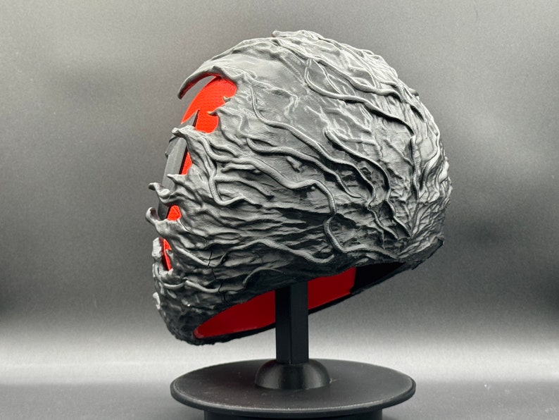 Puede incluir: Una m&aacute;scara negra impresa en 3D con un dise&ntilde;o texturizado y org&aacute;nico. La m&aacute;scara est&aacute; en un soporte negro y tiene un interior rojo.