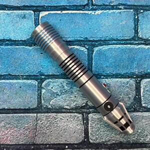 May include: A silver lightsaber prop with black and blue accents, featuring a ribbed handle and a detailed emitter. The prop has red and green buttons and is set against a blue brick wall background.