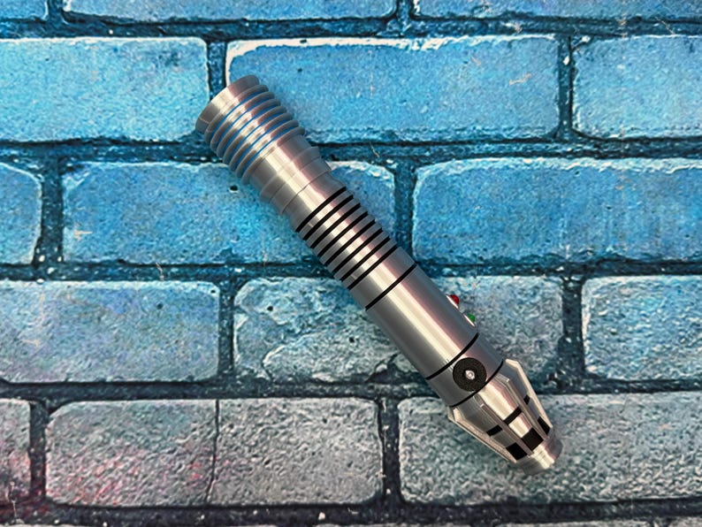 May include: A silver lightsaber prop with black and blue accents, featuring a ribbed handle and a detailed emitter. The prop has red and green buttons and is set against a blue brick wall background.