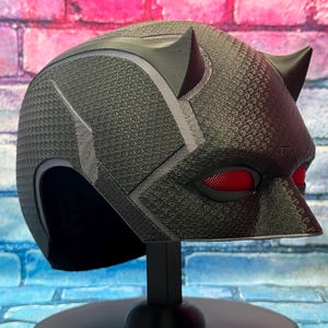 Puede incluir: Un casco negro con una superficie texturizada y lentes oculares rojas. El casco tiene dos cuernos puntiagudos en la parte superior y se exhibe sobre un soporte negro. El fondo es una colorida pared de ladrillos.