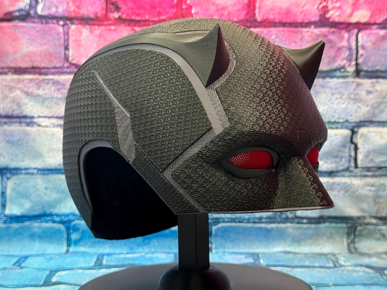 Puede incluir: Un casco negro con una superficie texturizada y lentes oculares rojas. El casco tiene dos cuernos puntiagudos en la parte superior y se exhibe sobre un soporte negro. El fondo es una colorida pared de ladrillos.