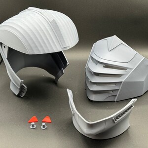 Puede incluir: Un casco gris impreso en 3D con una m&aacute;scara facial y una barbilla. El casco tiene una superficie texturizada y una visera. La m&aacute;scara facial tiene un dise&ntilde;o geom&eacute;trico con una serie de l&iacute;neas verticales. La barbilla es una pieza separada que se une al casco.