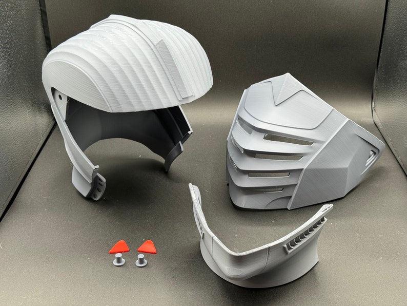 Puede incluir: Un casco gris impreso en 3D con una m&aacute;scara facial y una barbilla. El casco tiene una superficie texturizada y una visera. La m&aacute;scara facial tiene un dise&ntilde;o geom&eacute;trico con una serie de l&iacute;neas verticales. La barbilla es una pieza separada que se une al casco.