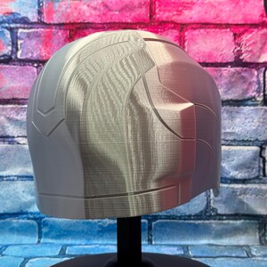 May include: A light gray, 3D-printed helmet with a textured surface. The helmet has a rounded top and angular side panels. It is displayed on a black stand against a brick wall with blue, pink, and purple paint.
