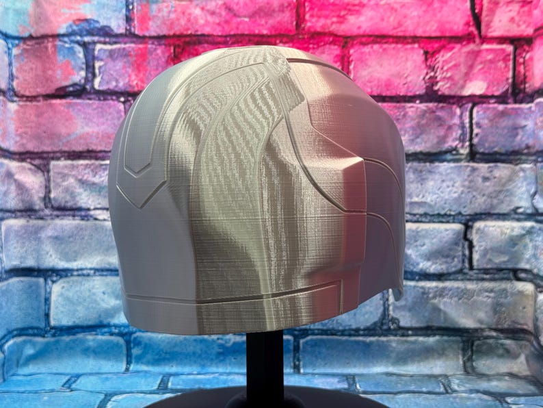 May include: A light gray, 3D-printed helmet with a textured surface. The helmet has a rounded top and angular side panels. It is displayed on a black stand against a brick wall with blue, pink, and purple paint.