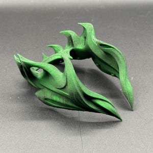 Puede incluir: Un objeto verde abstracto impreso en 3D con un dise&ntilde;o org&aacute;nico y fluido. El objeto tiene m&uacute;ltiples formas curvas en forma de hoja y una superficie texturizada. El color es un verde vibrante y s&oacute;lido, y el objeto parece estar sobre una superficie gris.