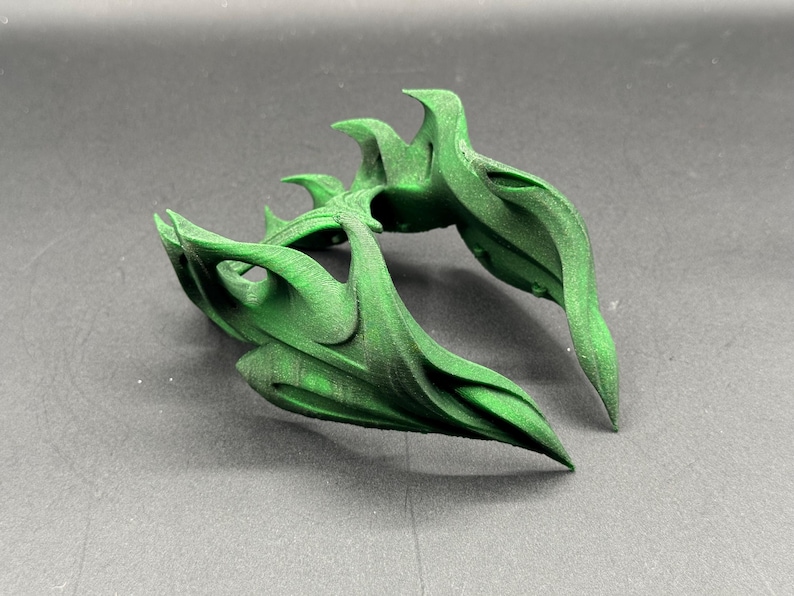 Puede incluir: Un objeto verde abstracto impreso en 3D con un dise&ntilde;o org&aacute;nico y fluido. El objeto tiene m&uacute;ltiples formas curvas en forma de hoja y una superficie texturizada. El color es un verde vibrante y s&oacute;lido, y el objeto parece estar sobre una superficie gris.