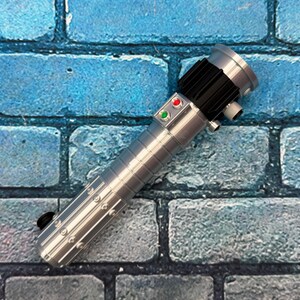 Op de afbeelding: Een zilveren lichtzwaard prop met een zwarte grip en accenten. De prop heeft rode en groene knoppen en staat tegen een blauwe bakstenen muur achtergrond. Het lichtzwaard heeft een gedetailleerd, metalen ontwerp.