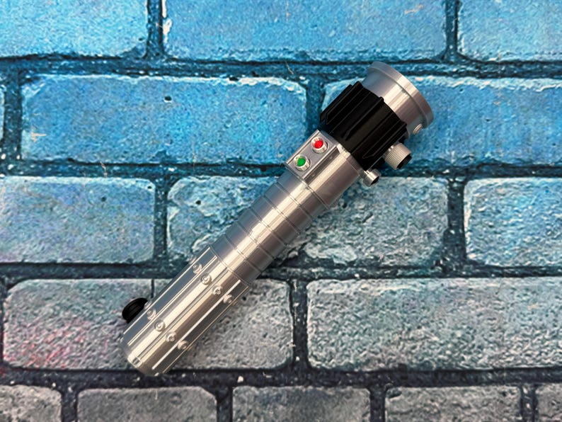 Op de afbeelding: Een zilveren lichtzwaard prop met een zwarte grip en accenten. De prop heeft rode en groene knoppen en staat tegen een blauwe bakstenen muur achtergrond. Het lichtzwaard heeft een gedetailleerd, metalen ontwerp.