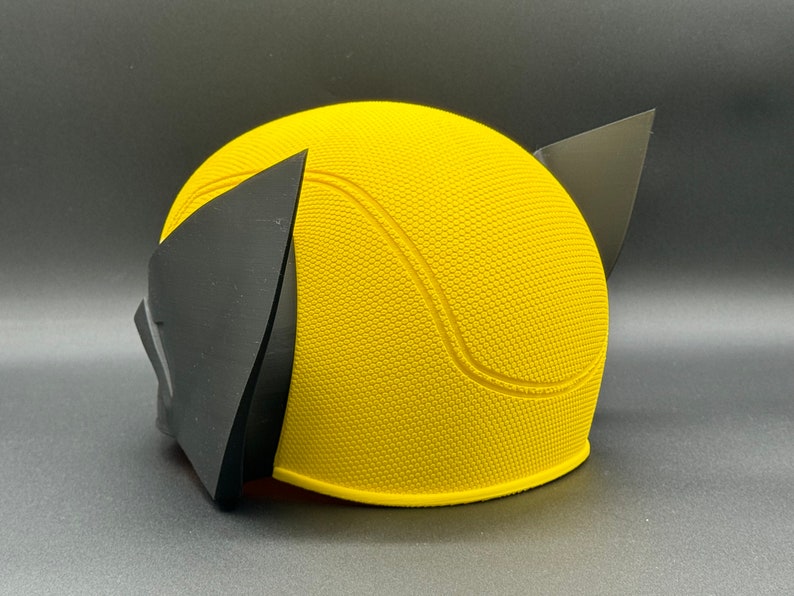 May include: Yellow and black plastic costume helmet with a textured surface. The helmet has a rounded shape with a pointed top and two black ear pieces.