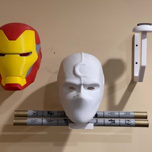 Może przedstawiać: Prezentacja na ścianie przedmiot&oacute;w o tematyce superbohaterskiej, w tym maska Iron Mana w kolorze czerwonym, ż&oacute;łtym i szarym, biała maska Moon Knight i dwa cylindryczne przedmioty z szarymi i złotymi akcentami. Obecny jest r&oacute;wnież biały uchwyt na kask.