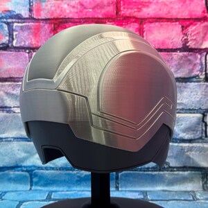 May include: A futuristic-style helmet with a silver and gray color scheme. The helmet has a smooth, rounded design with angular accents and a visor area. It is displayed on a black stand against a brick wall background.