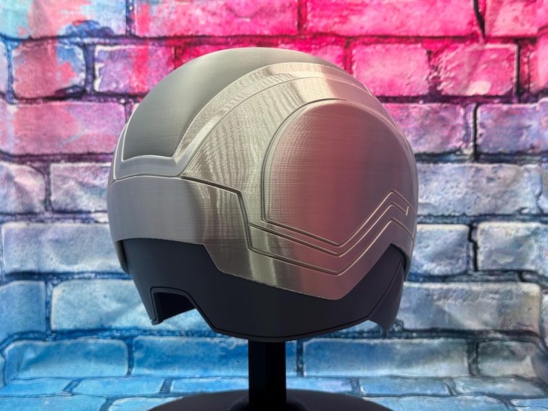 May include: A futuristic-style helmet with a silver and gray color scheme. The helmet has a smooth, rounded design with angular accents and a visor area. It is displayed on a black stand against a brick wall background.
