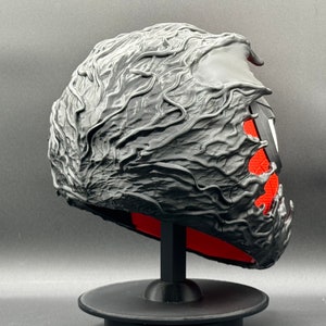 Puede incluir: Una m&aacute;scara negra impresa en 3D con un dise&ntilde;o texturizado y org&aacute;nico. La m&aacute;scara est&aacute; en un soporte negro con una base roja.