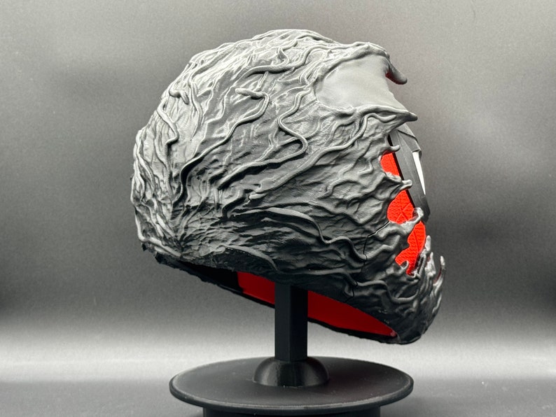 Puede incluir: Una m&aacute;scara negra impresa en 3D con un dise&ntilde;o texturizado y org&aacute;nico. La m&aacute;scara est&aacute; en un soporte negro con una base roja.