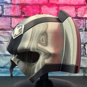 May include: A silver and black helmet with a distinctive design, featuring a crest emblem on the side. The helmet has a metallic sheen and a sleek, modern aesthetic. The helmet is displayed against a colorful brick wall.