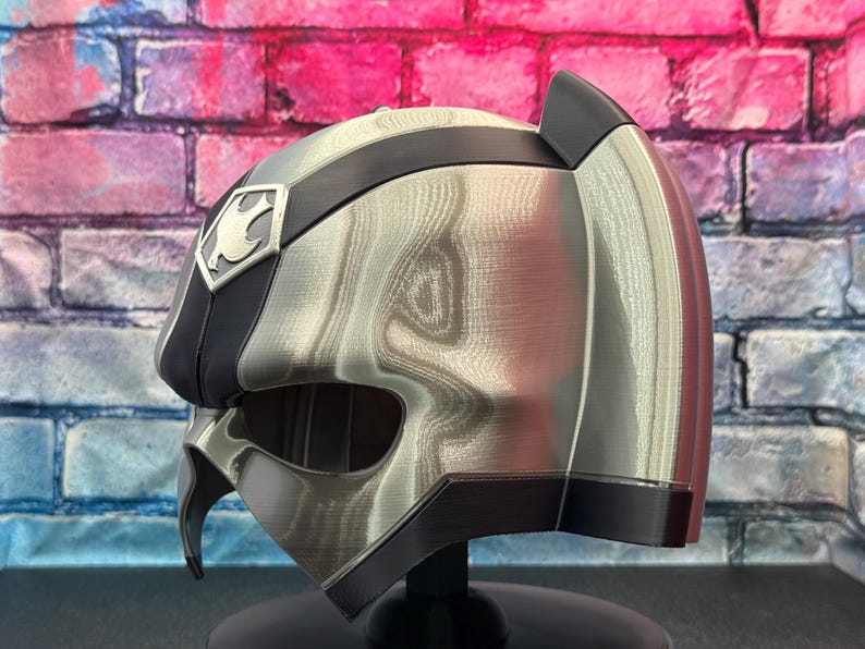 May include: A silver and black helmet with a distinctive design, featuring a crest emblem on the side. The helmet has a metallic sheen and a sleek, modern aesthetic. The helmet is displayed against a colorful brick wall.