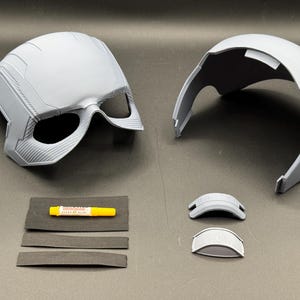 May include: A disassembled gray helmet with eye openings, alongside a separate top piece, two small visor components, and adhesive. The helmet has detailed surface textures and appears to be 3D-printed. Accessories include foam strips.