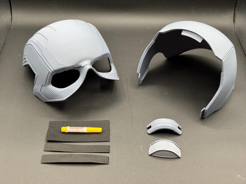 May include: A disassembled gray helmet with eye openings, alongside a separate top piece, two small visor components, and adhesive. The helmet has detailed surface textures and appears to be 3D-printed. Accessories include foam strips.