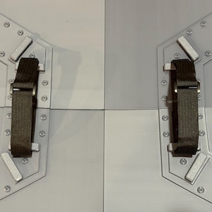 May include: Two light gray, segmented panels with dark brown straps and silver-colored hardware. The panels are connected by a seam and feature a series of small, silver rivets around the edges. The straps are attached to the panels with silver hardware.