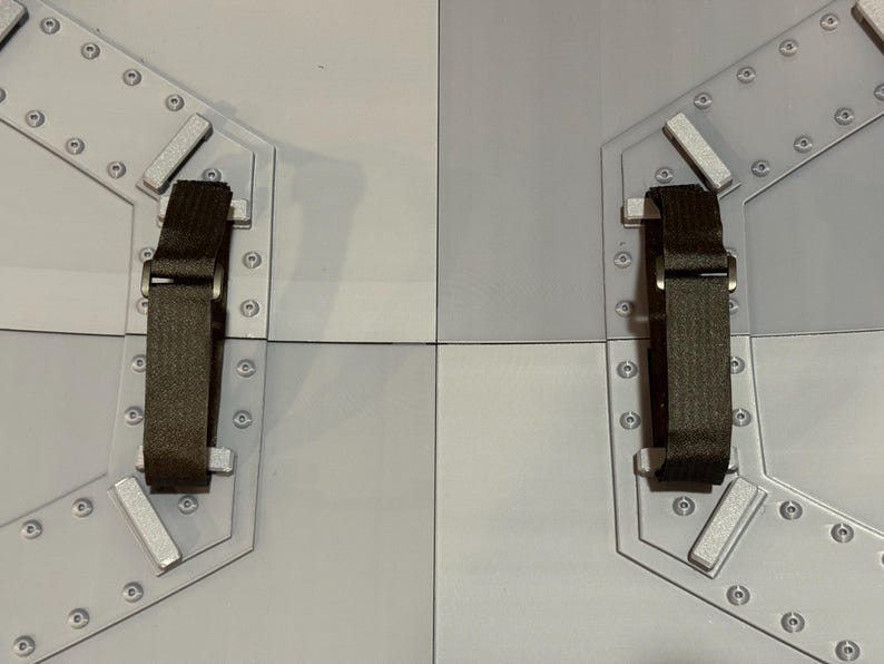 May include: Two light gray, segmented panels with dark brown straps and silver-colored hardware. The panels are connected by a seam and feature a series of small, silver rivets around the edges. The straps are attached to the panels with silver hardware.