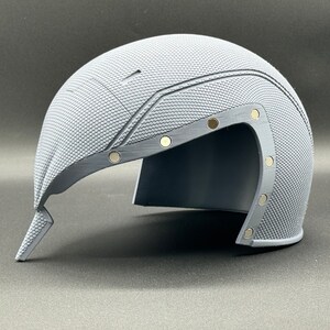 May include: A gray helmet with a textured surface and a visor. The helmet has several small, round, silver-colored accents.
