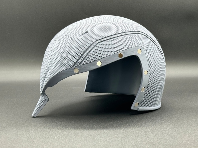 May include: A gray helmet with a textured surface and a visor. The helmet has several small, round, silver-colored accents.