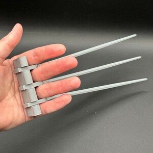 May include: A silver-colored, three-fingered claw accessory with three retractable blades. The claws are worn on the hand, with the blades extending out from the fingers.