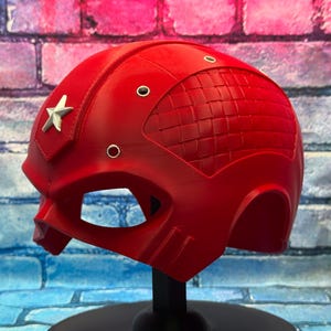 May include: A bright red helmet with a silver star emblem on the side. The helmet has a textured design on the top and several small circular details. It is displayed on a black stand against a brick wall.