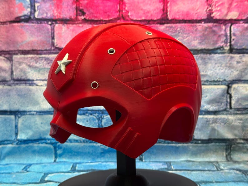 May include: A bright red helmet with a silver star emblem on the side. The helmet has a textured design on the top and several small circular details. It is displayed on a black stand against a brick wall.
