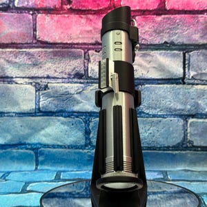 May include: A silver and black lightsaber prop, standing upright. The hilt is silver with black accents, and the top features a black emitter. The lightsaber is displayed on a black stand against a brick wall.