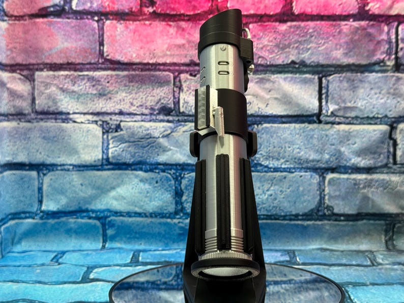 May include: A silver and black lightsaber prop, standing upright. The hilt is silver with black accents, and the top features a black emitter. The lightsaber is displayed on a black stand against a brick wall.