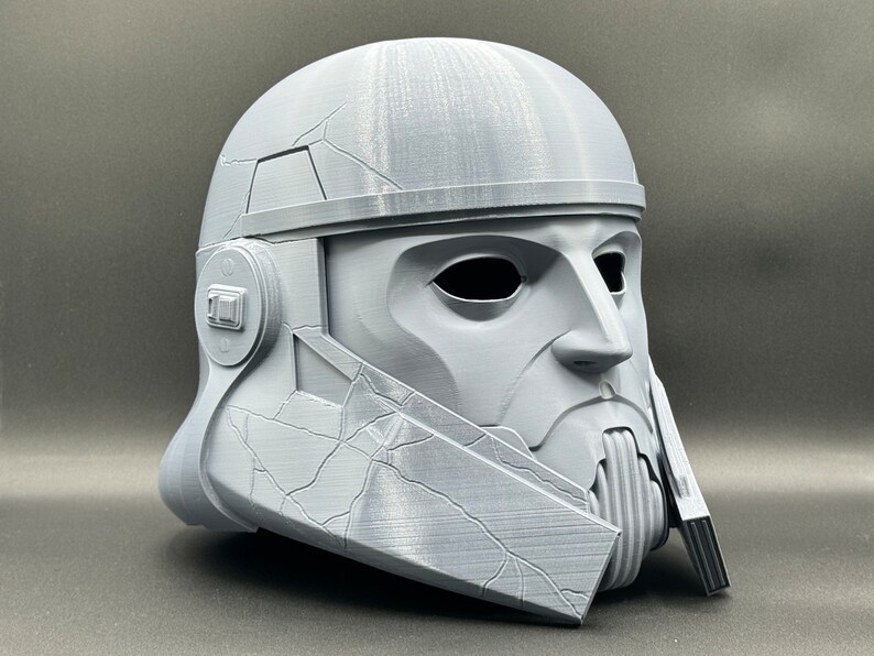 May include: A gray 3D printed helmet with a textured surface and a detailed face mask. The helmet has a raised ridge around the top and a small, round detail on the side.