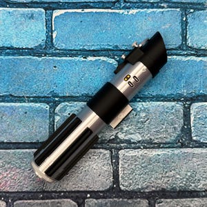 May include: A silver and black lightsaber prop with a black handle and a ribbed lower section. The prop has a black emitter and several silver buttons and details. It rests on a blue brick background.