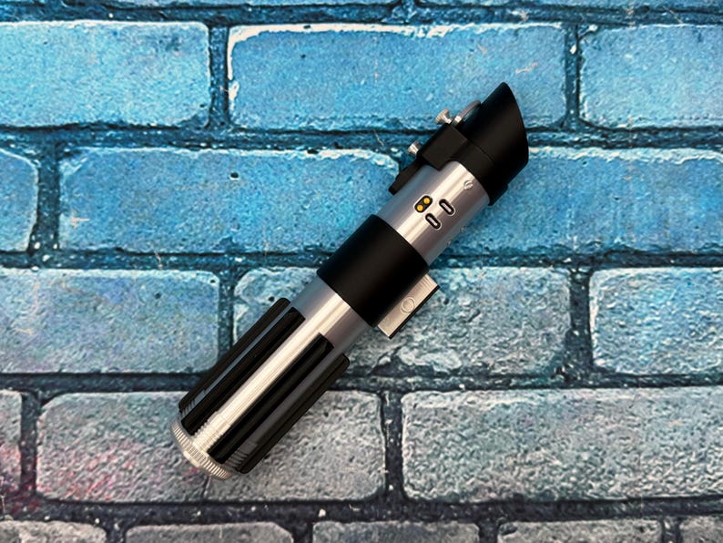 May include: A silver and black lightsaber prop with a black handle and a ribbed lower section. The prop has a black emitter and several silver buttons and details. It rests on a blue brick background.